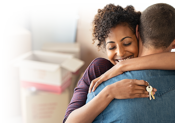 Photo of a young woman with a set of house keys hugging a young man with moving boxes in the background.