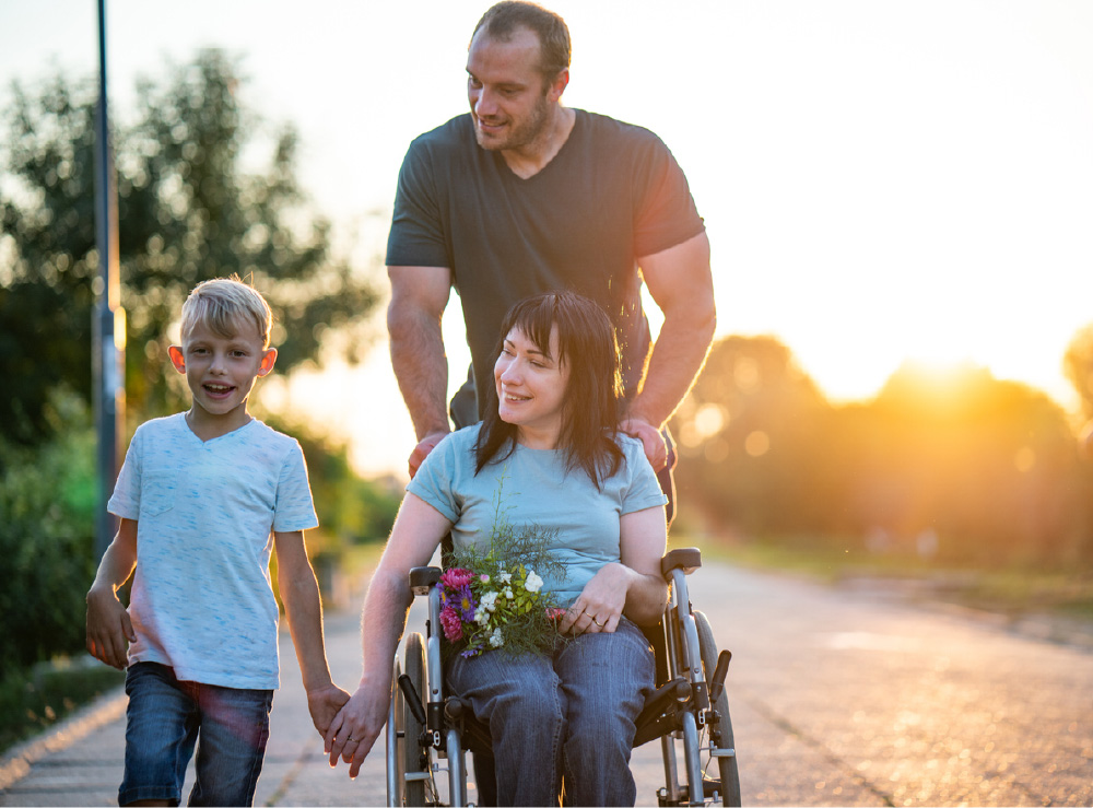Photo of young family on an evening walk with young boy and father behind mother in wheelchair. Photo of young family on an evening walk with young boy and father behind mother in wheelchair.